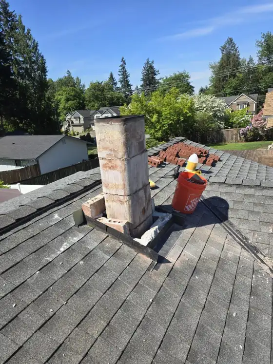 Wide view of a chimney being rebuilt, with a bucket and new bricks on the shingle roof