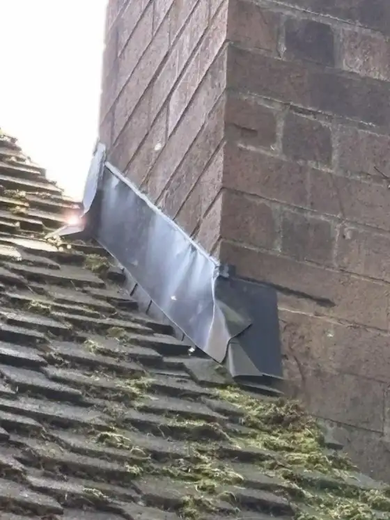 Metal flashing lifting at the base of a brick chimney with moss on the shingles