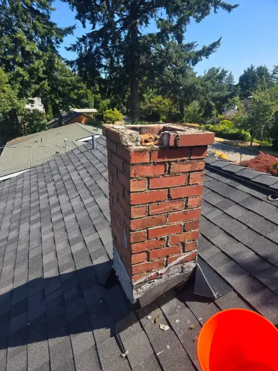 Narrow brick chimney on a shingle roof with a broken, crumbling top and an orange bucket beside it, before repair