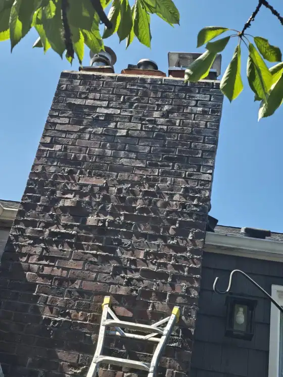 Tall brick chimney dark with soot and staining on the side of a gray house, with a ladder against it, before cleaning