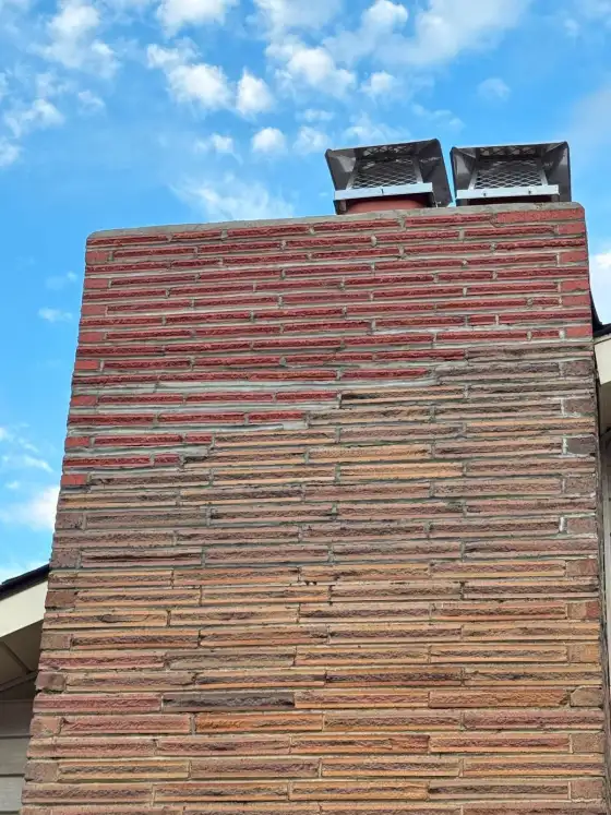 Tall chimney faced with long thin tan and red bricks, topped with two new mesh caps against a blue sky