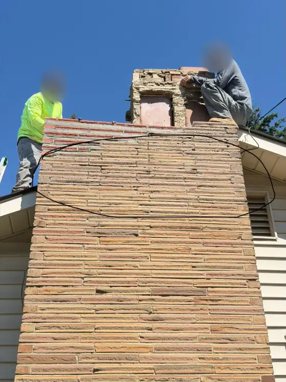 Two masons working on the crumbling top of a tall thin-brick chimney against a blue sky, faces blurred